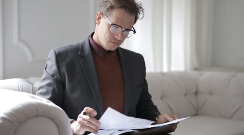 Elegant adult man in jacket and glasses looking through documents while sitting on white sofa in luxury room