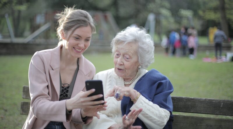 Delighted female relatives sitting together on wooden bench in park and browsing mobile phone while learning using