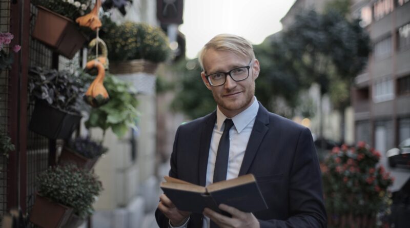 Confident male manager in glasses holding book and looking at camera while standing near plant store on sidewalk in city