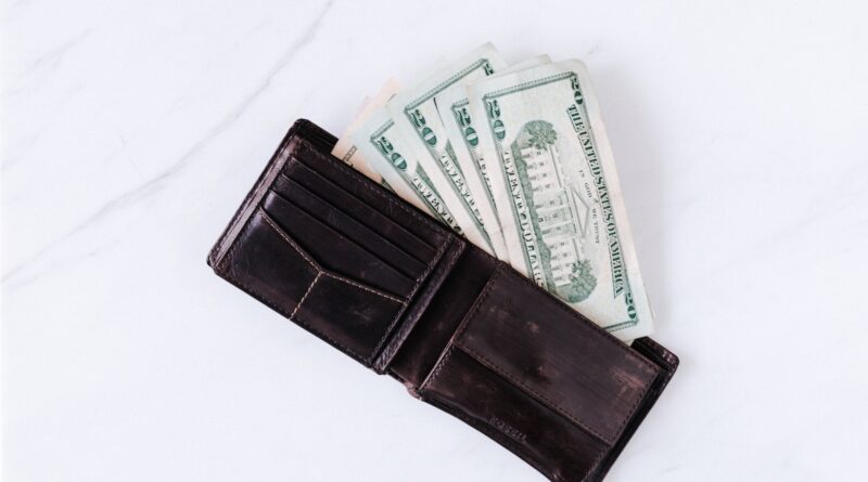From above of black leather wallet with pockets filled with heap of dollar banknotes on white table