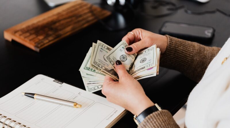 Unrecognizable elegant female in sweater counting dollar bills while sitting at wooden table with planner and pen