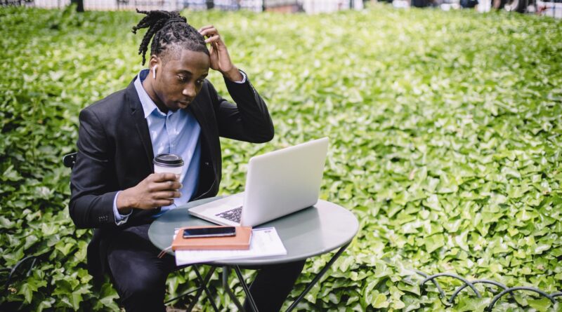 Focused young African American male freelancer with dreadlocks in formal wear drinking coffee in street cafe and scratching head while reading report on laptop