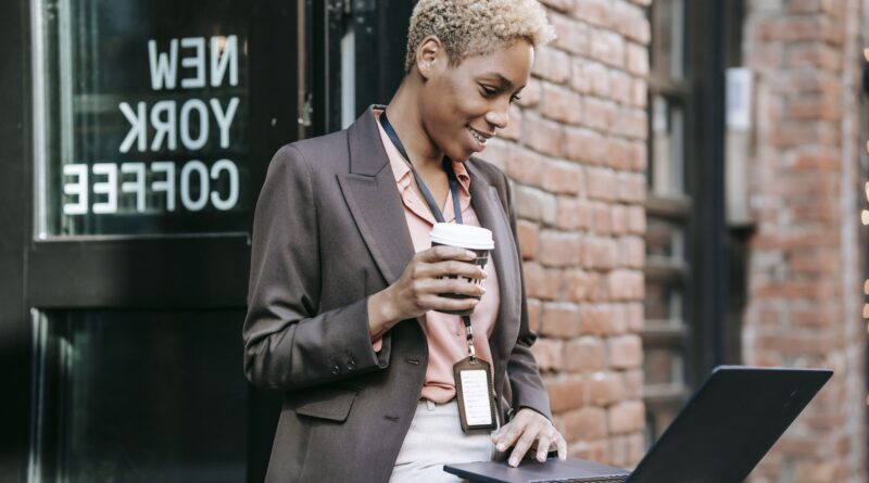 Positive African American businesswoman with cup of coffee browsing netbook in outdoor cafeteria while working remotely
