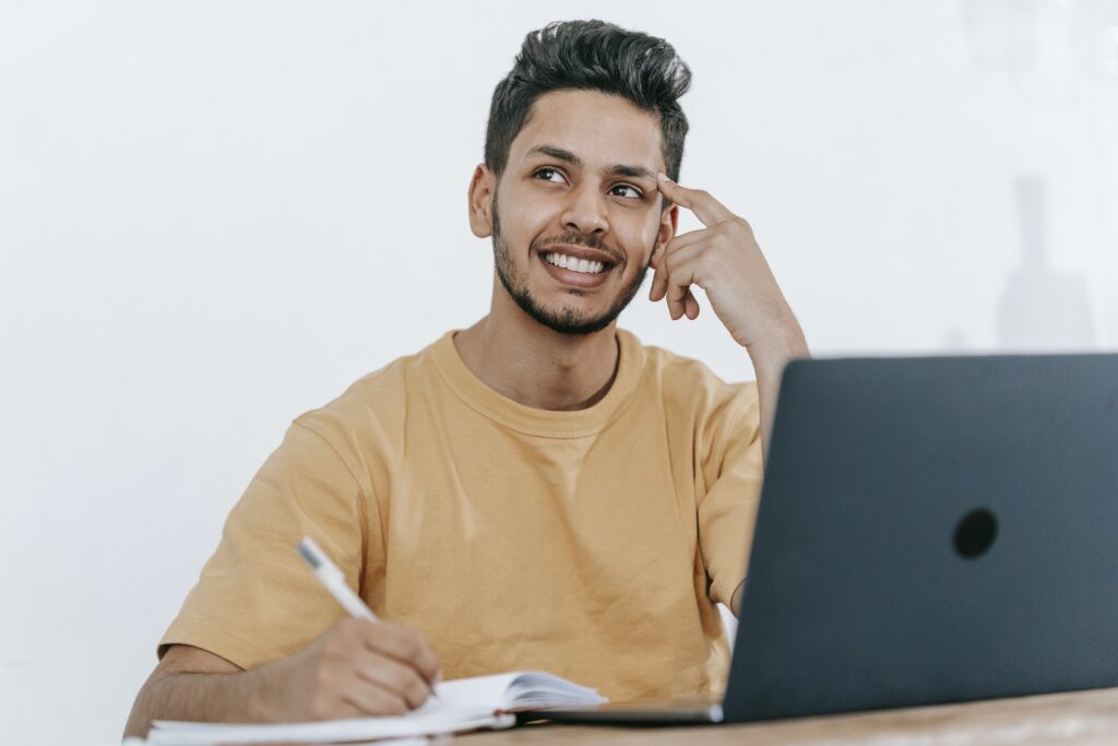 Smiling young bearded Hispanic male entrepreneur thinking over new ideas for startup project and looking away dreamily while working at table with laptop and taking notes in notebook