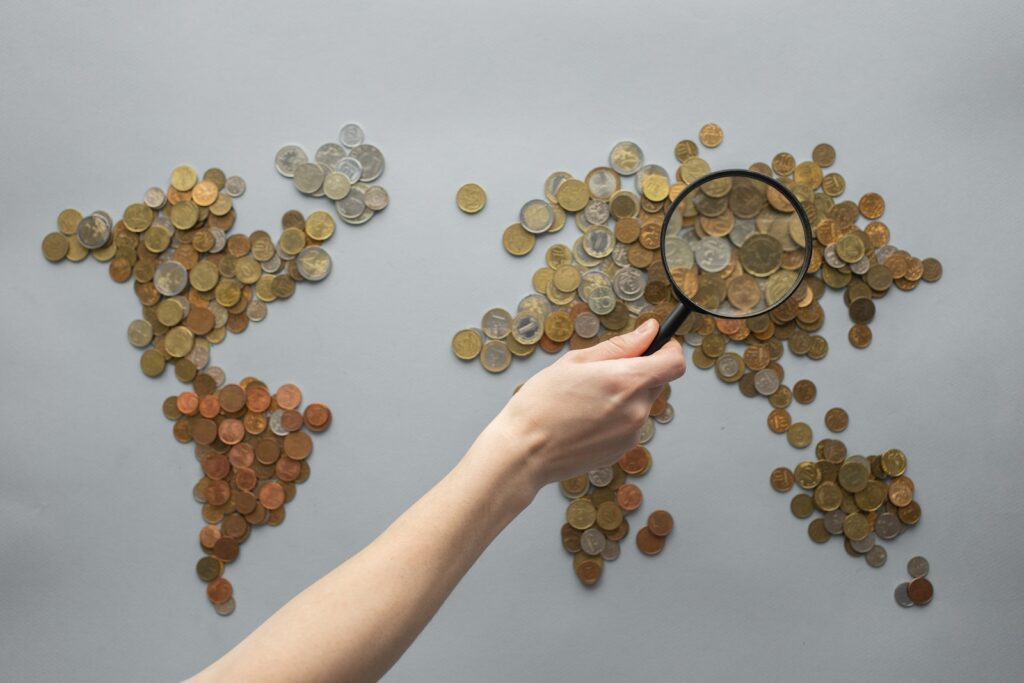 Top view of crop unrecognizable traveler with magnifying glass standing over world map made of various coins on gray background